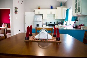 A dining area with a wooden table, chairs, and a placemat. There is a kitchen in the background with white cabinets, a refrigerator, and a stove.