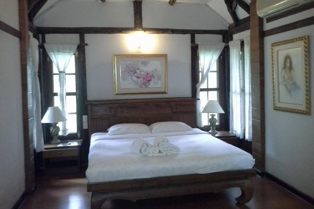 Una habitación tradicional con una cama de madera, dos mesitas de noche con lámparas, un cuadro enmarcado en la pared y una ventana con cortinas.