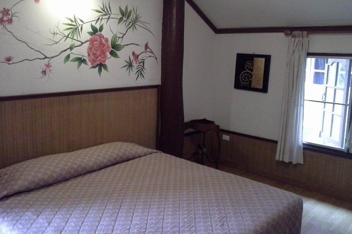 Un dormitorio con una cama, una pared decorada con motivos florales, una ventana con cortinas y una mesita.
