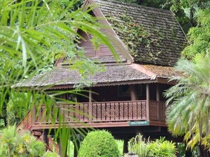 A traditional wooden house with a thatched roof, surrounded by lush greenery and palm trees. - Baanklangdoi Resort (Hang Dong)