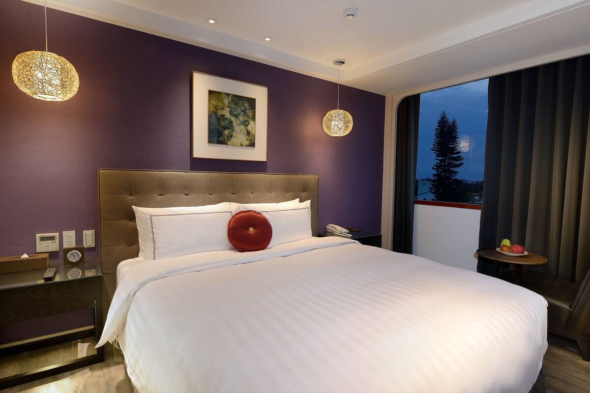 A hotel room with a large bed, a bedside table with a phone, and a view of a tree through the window.