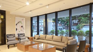 A modern hotel lobby with a beige sofa, two armchairs, and a glass coffee table. Large windows offer a view of the street and greenery outside. - Shiny Ocean Hotel (Hualien City)