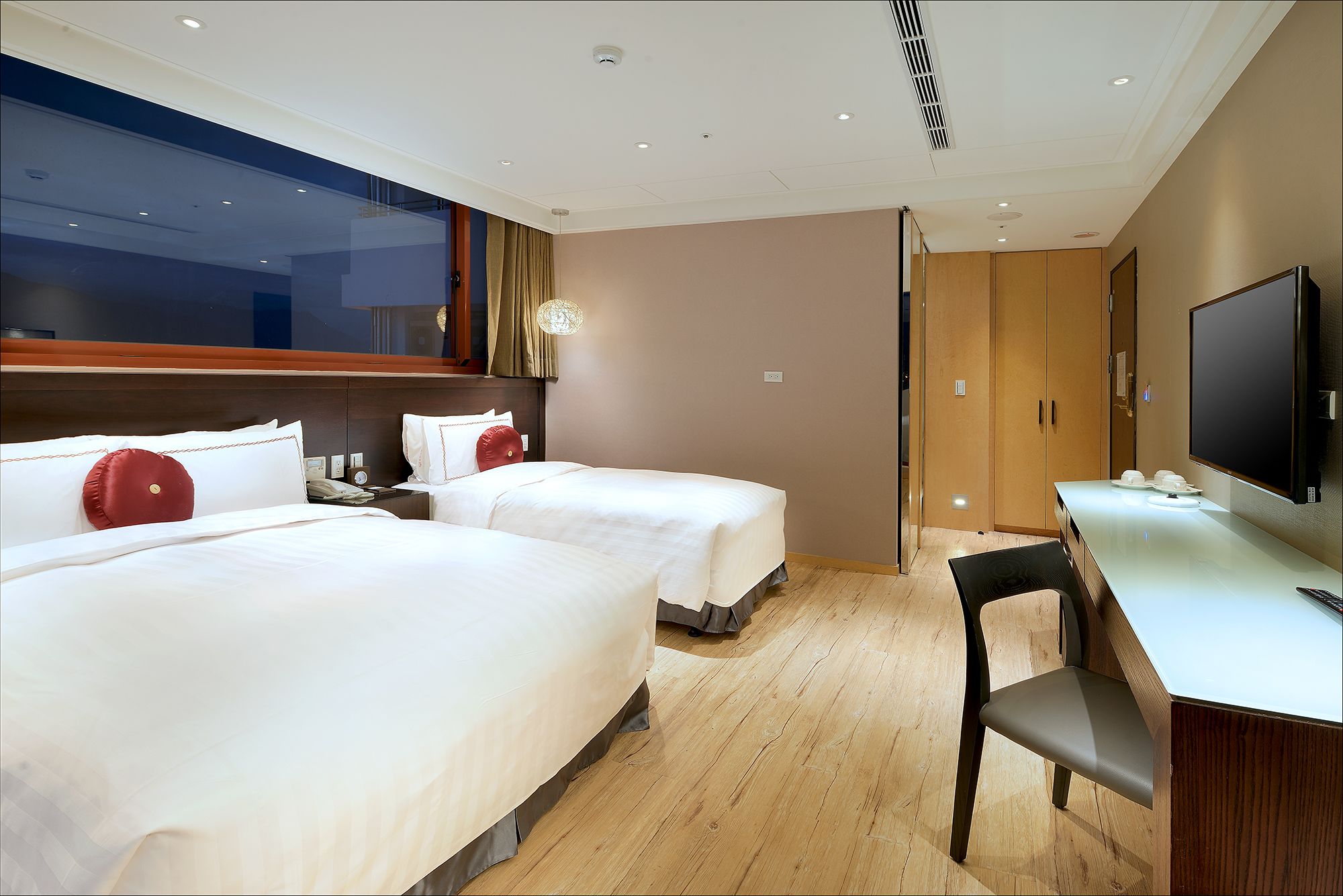 Una habitación de hotel con dos camas, un escritorio con silla, un televisor y un ventanal amplio.