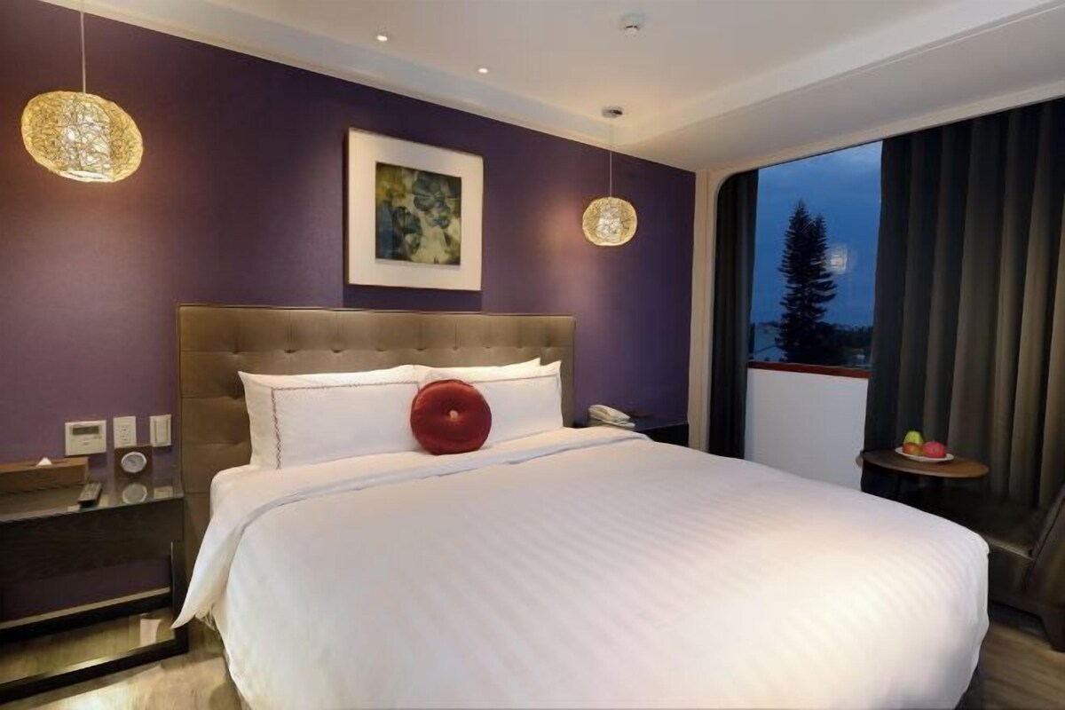 A modern hotel room with a large bed, a nightstand, a painting on the wall, and a view of the outdoors.