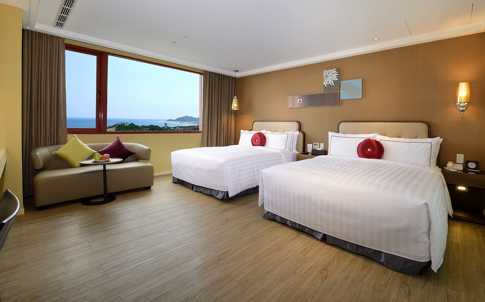 A hotel room with two beds, a sofa, a small table, and a view of the sea.