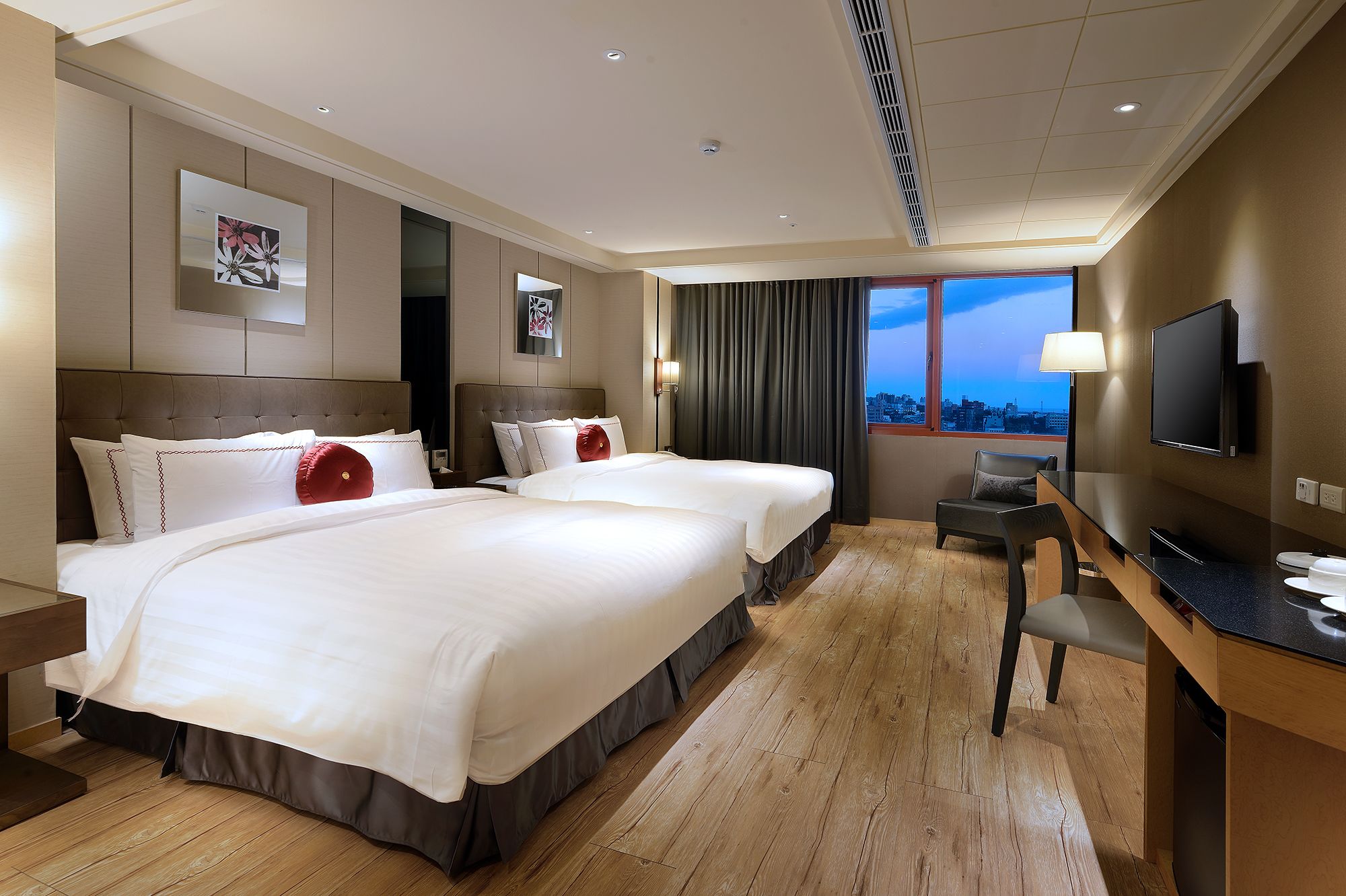 A hotel room with two beds, a desk, a chair, a TV, and a city view.