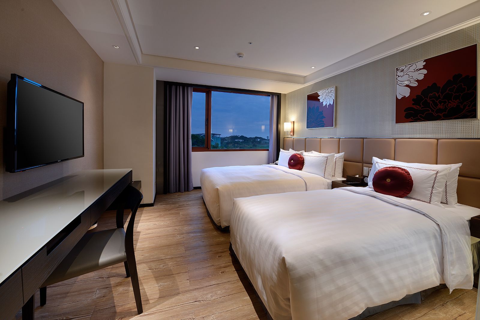 A hotel room with two beds, a desk, a TV, and a large window with a city view.