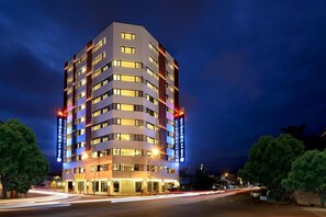 Un hotel de varios pisos con ventanas iluminadas y señalización, fachada plana, y vista a la calle con estelas de luz.