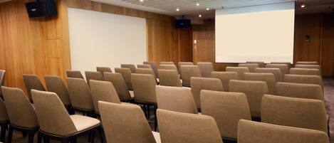 Un salón de conferencias con filas de sillas, una pantalla para proyector y una pantalla grande.