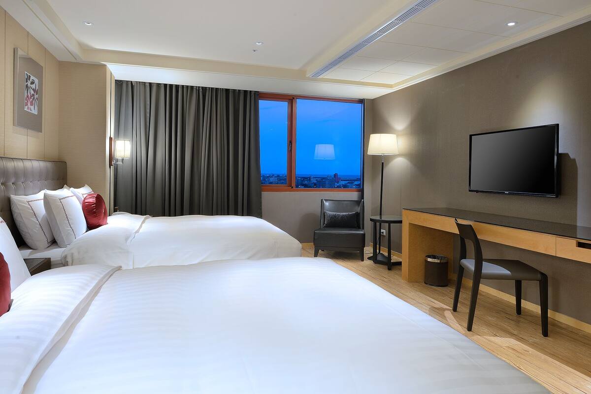A hotel room with two beds, a desk, a chair, a TV, and a window with curtains.
