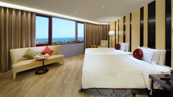 A hotel room with a large bed, a sofa, a small table, and a view of the sea through large windows.