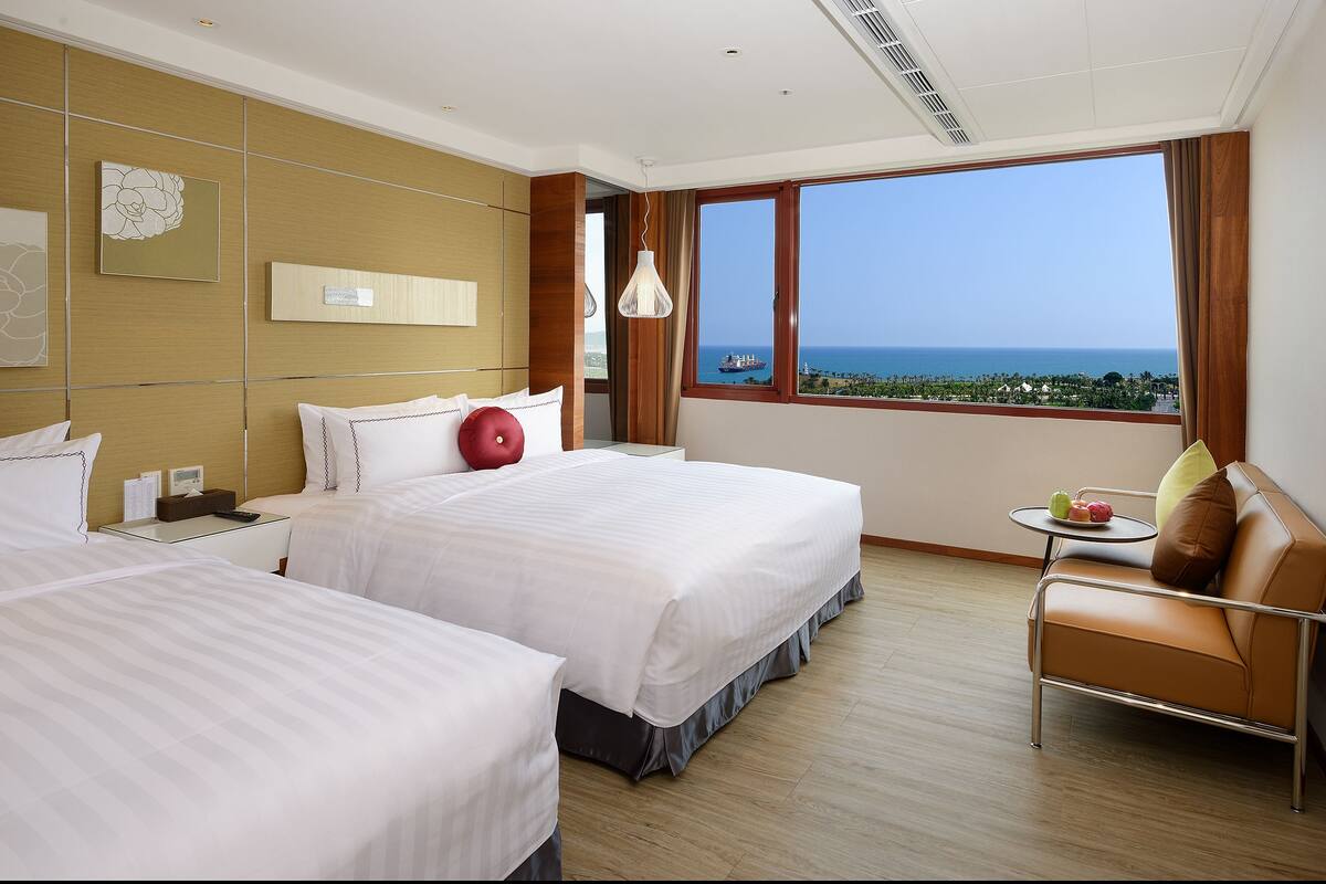 A hotel room with two beds, a sofa, a small table with fruit, and a large window with a view of the sea.