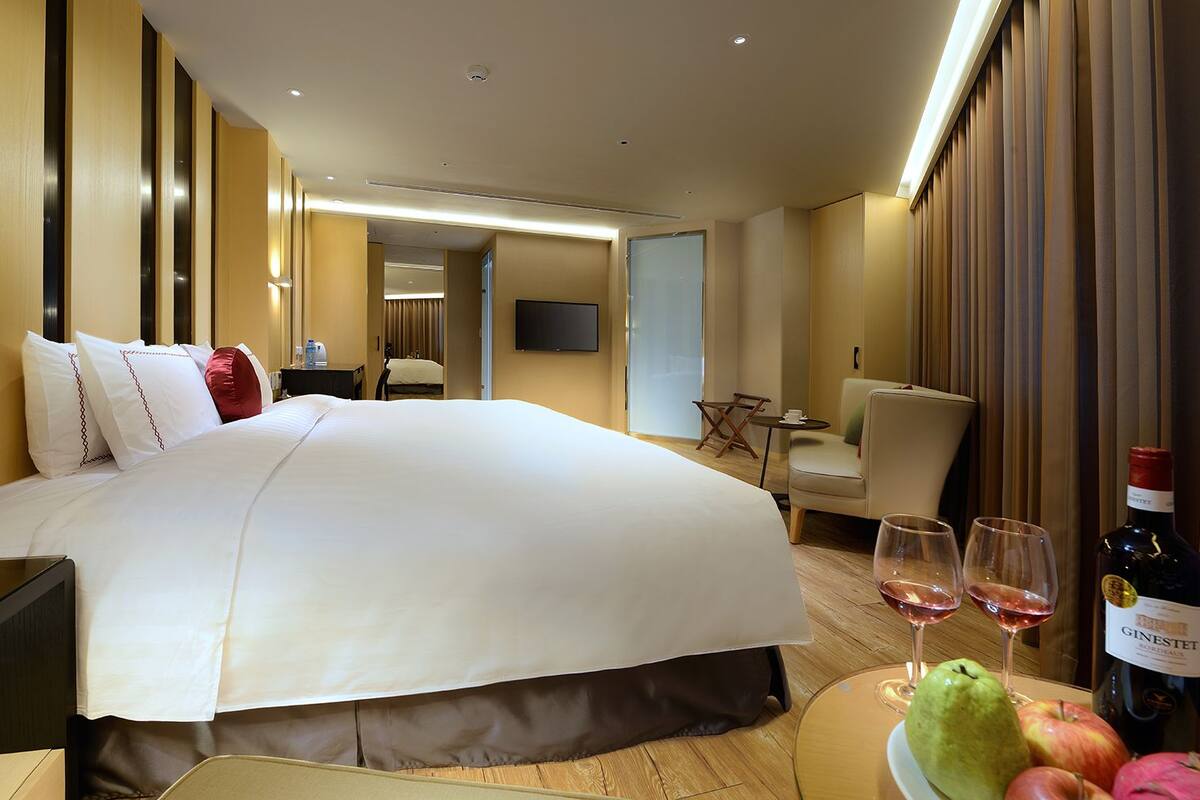 A hotel room with a large bed, a bedside table, a chair, a small table with wine glasses and fruit, and a television on the wall.