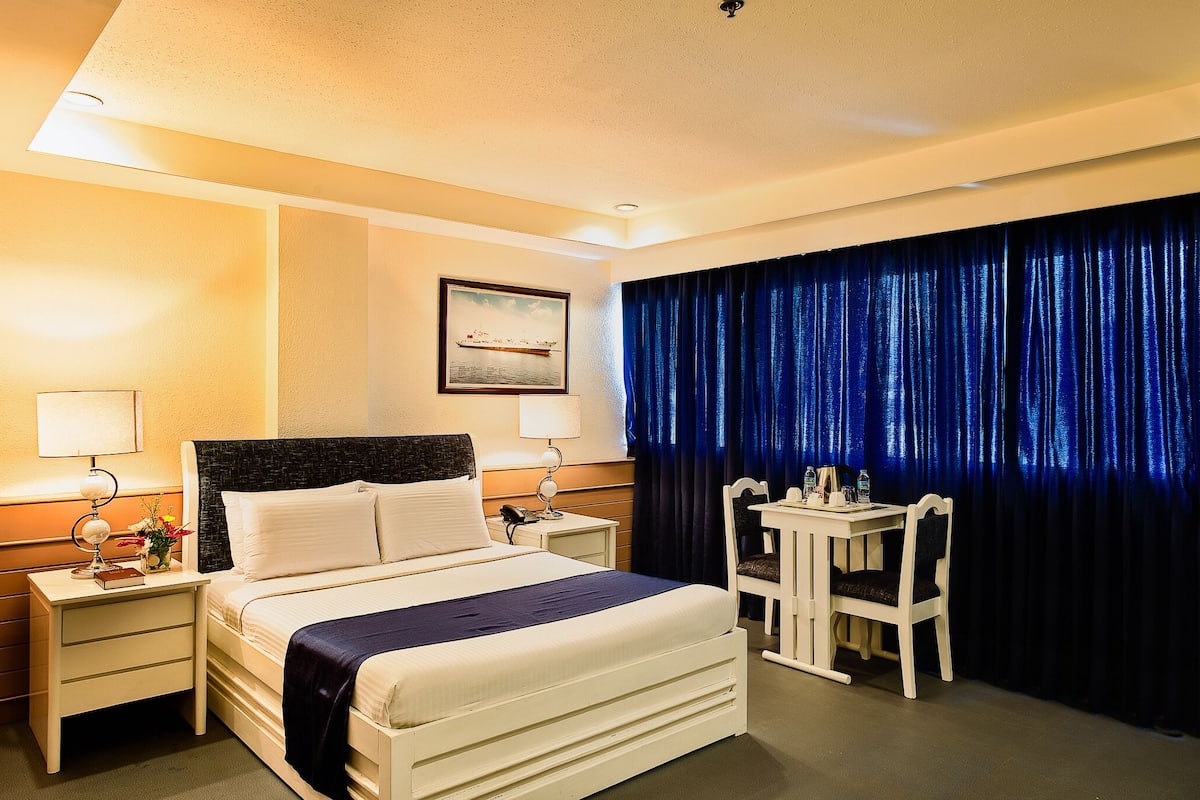 Ein Hotelzimmer mit Bett, Nachttischen, einem Schreibtisch mit Stuhl, einem Fenster mit Vorhängen und einem Bild an der Wand.
