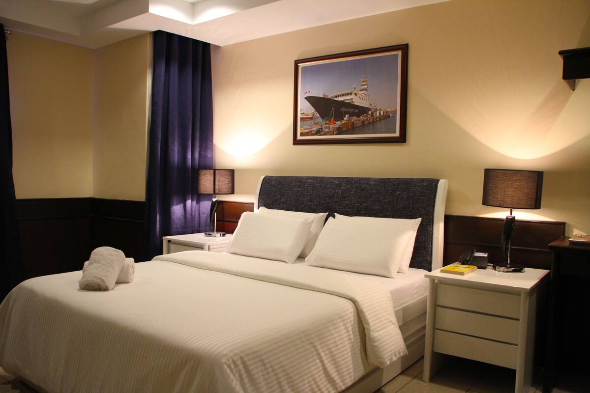 Ein Hotelzimmer mit einem großen Bett, zwei Nachttischen, einer Lampe, einem gerahmten Bild an der Wand und einem Vorhang.