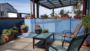 Uma área de varanda coberta com uma mesa e cadeiras de vidro, cercada por vasos de plantas e uma parede azul.