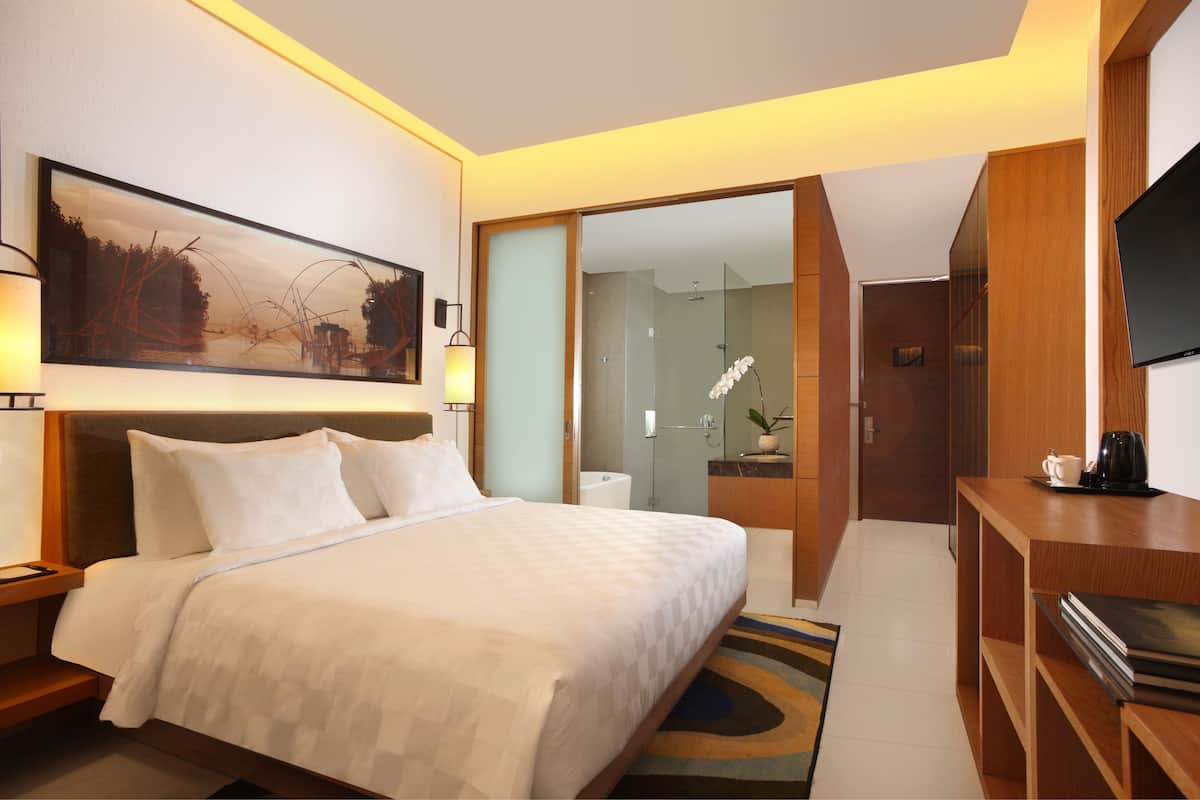 A hotel room with a large bed, a TV, a desk, and a bathroom visible through a glass door.