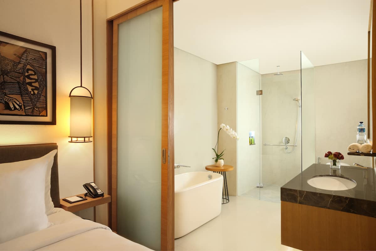 A modern hotel bathroom with a bathtub, a glass-enclosed shower, a vanity with a marble countertop, and a bedside table with a lamp and phone.