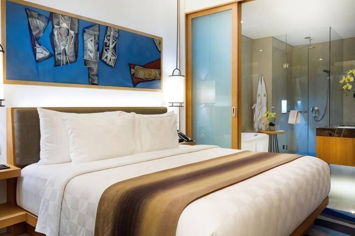 A hotel room with a bed, a shower, a glass-enclosed bathroom, and a painting on the wall.