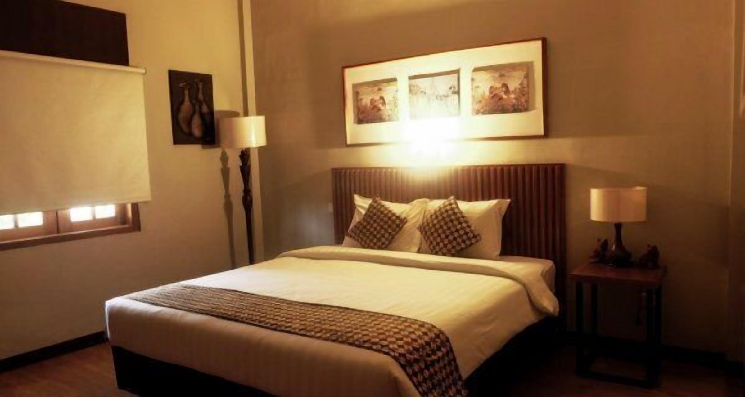 A hotel room with a large bed, a bedside table with a lamp, a window with blinds, and artwork on the wall.