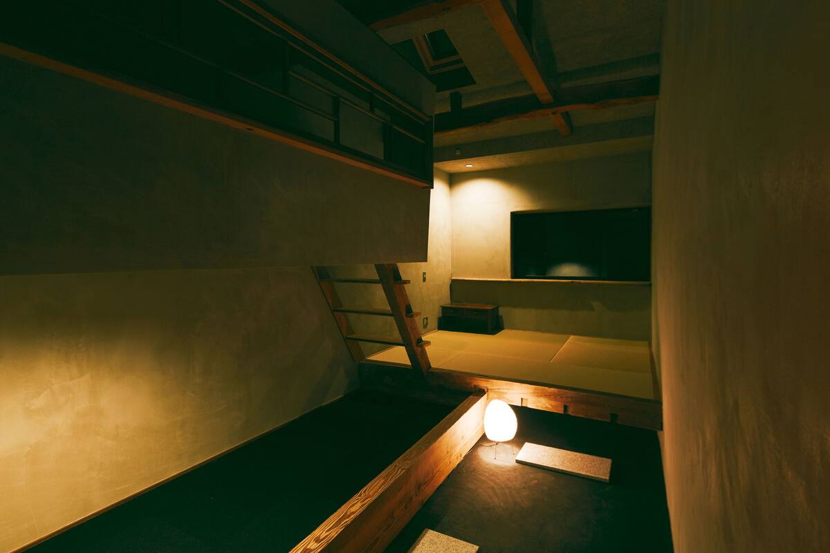 A dimly lit room with bunk beds, a ladder, and a small table with a light.