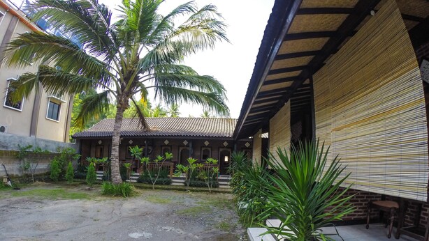 A courtyard with a palm tree, a covered seating area, and a building with a tiled roof. - Warungku In Home Stay Senggigi (Senggigi)