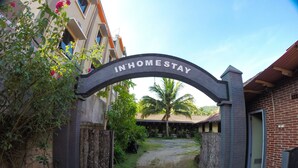 An archway with "IN-HOME STAY" written on it, leading to a courtyard with a palm tree and a building with a tiled roof. - Warungku In Home Stay Senggigi (Senggigi)