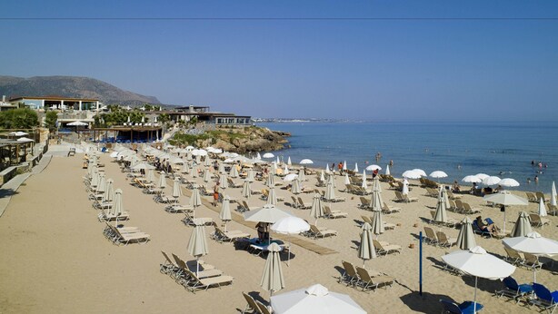 A beach with numerous sun loungers and umbrellas, a building with a sign reading "KAROS", and a clear view of the sea. - Arkadi Apartments by Checkin (Hersonissos)
