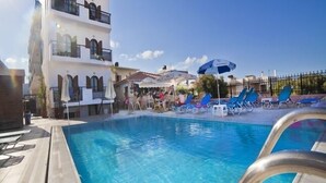 A swimming pool with lounge chairs and umbrellas, a multi-story building with balconies, and a clear blue sky. - Arkadi Apartments by Checkin (Hersonissos)