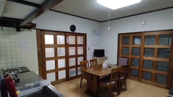 A room with a wooden dining table, chairs, a television, and a kitchen area with a refrigerator and sink.