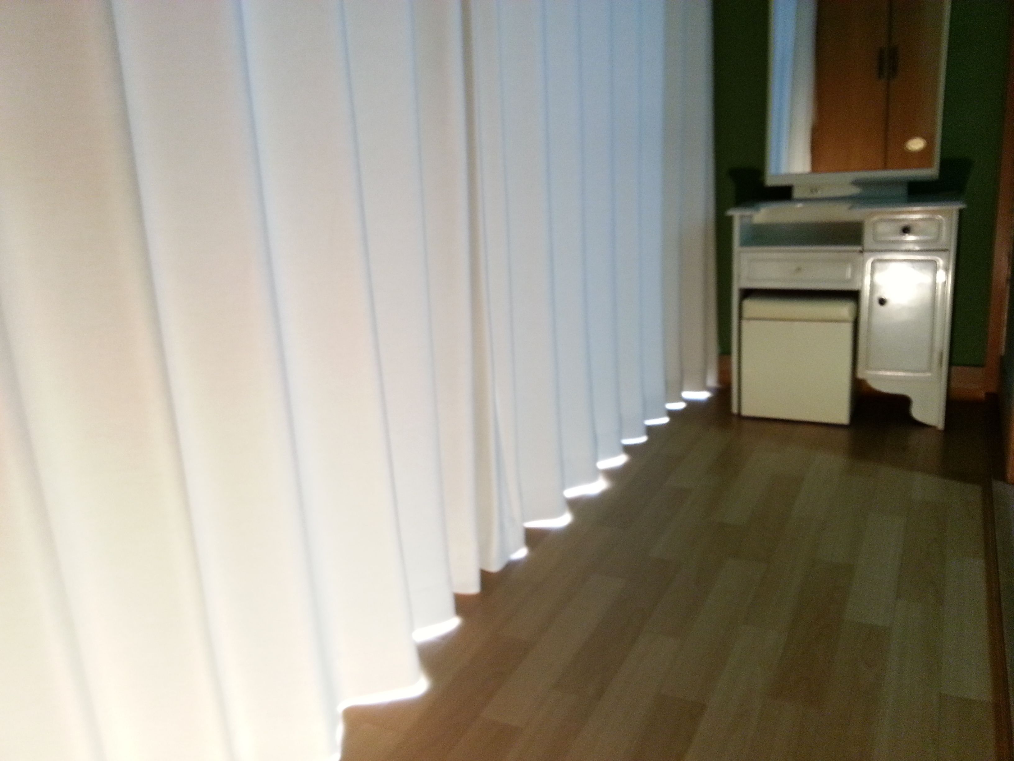 Een smalle gang met een houten vloer, witte gordijnen en een klein keukentje met een koelkast en een magnetron.