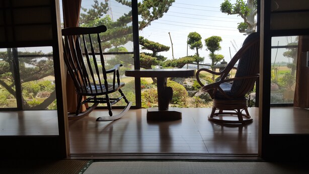 A room with two rocking chairs and a small table, large windows with a view of a garden and trees. - Guest House Nakamura House (Oki)