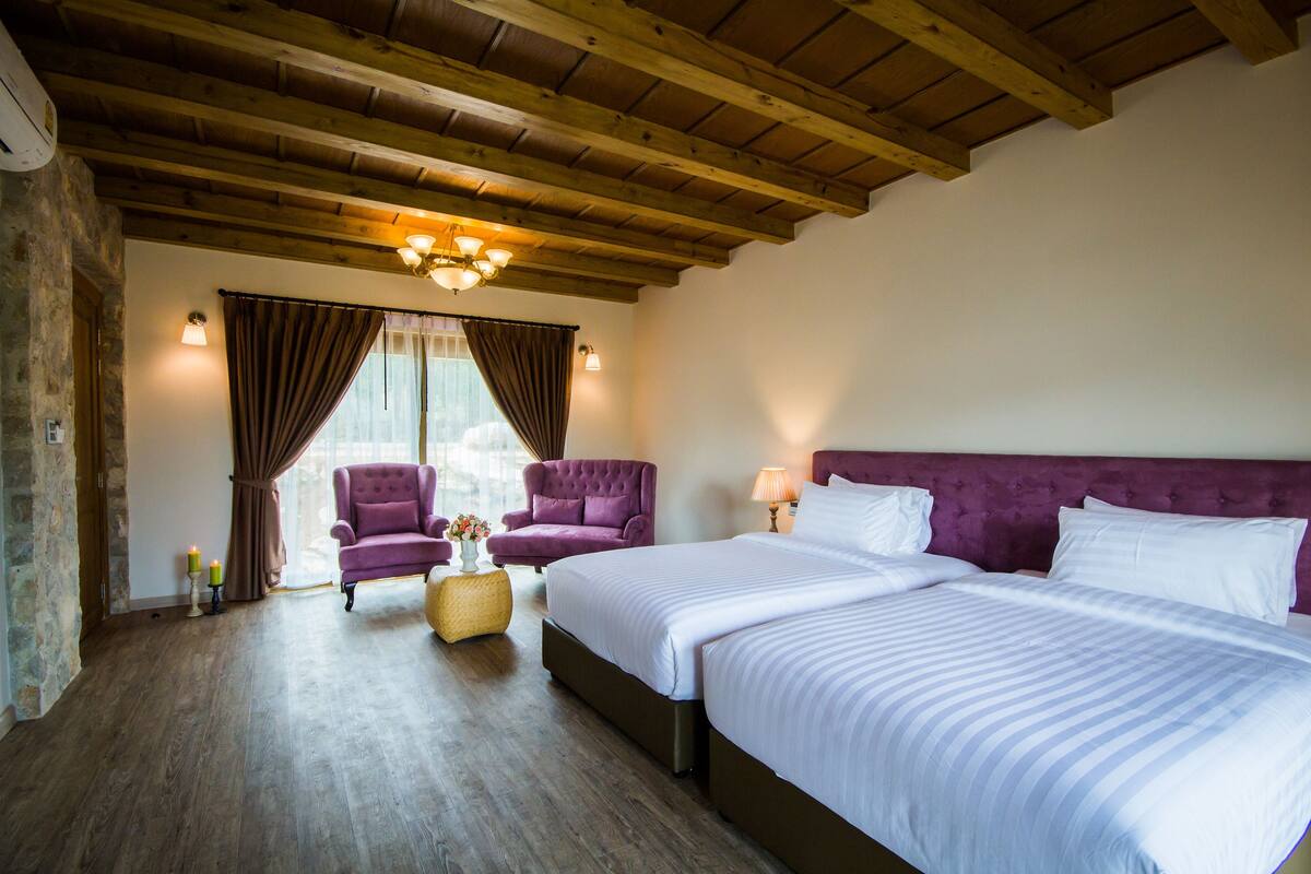 A hotel room with a wooden ceiling, two beds, a purple armchair, a small round table, and a window with curtains.