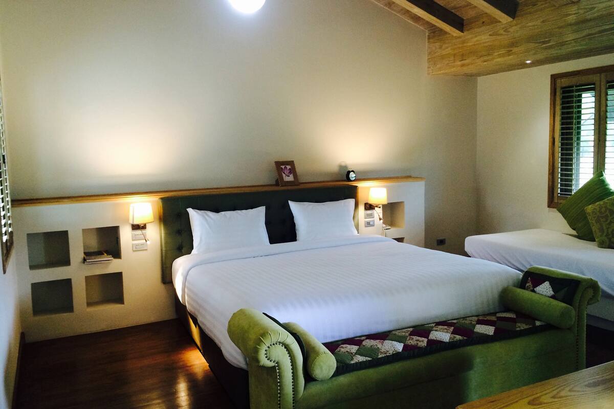 A hotel room with a large bed, a smaller bed, a green armchair, and a nightstand with a lamp.