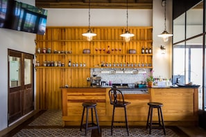 Una zona de bar con barra de madera, taburetes, un televisor montado en la pared y estantes con botellas y frascos.