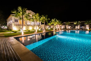 Un área de piscina nocturna con un edificio moderno, palmeras y sillones de descanso.