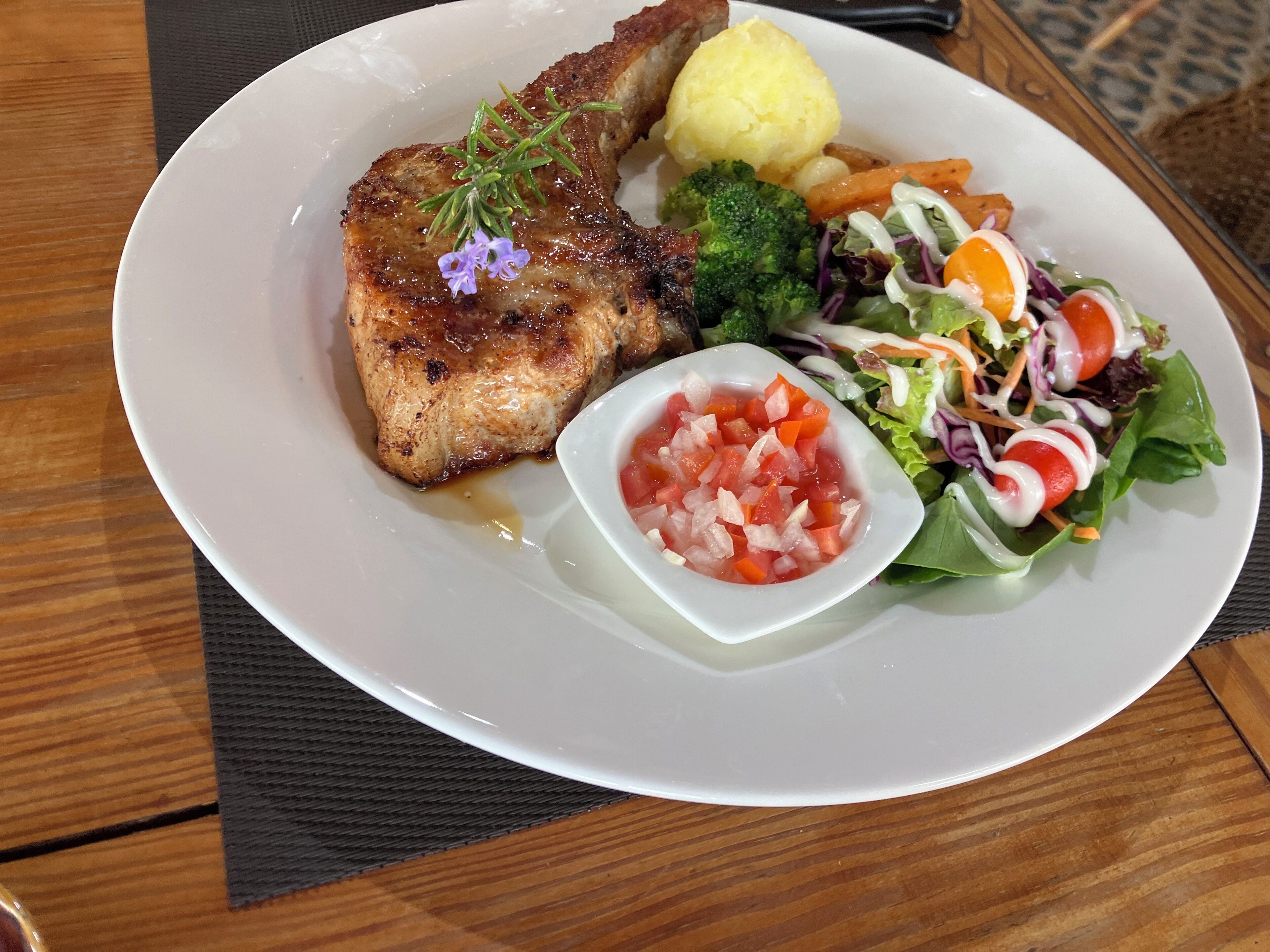 Un plato con carne a la parrilla, puré de papas, verduras al vapor y una ensalada con un recipiente pequeño de tomates picados.