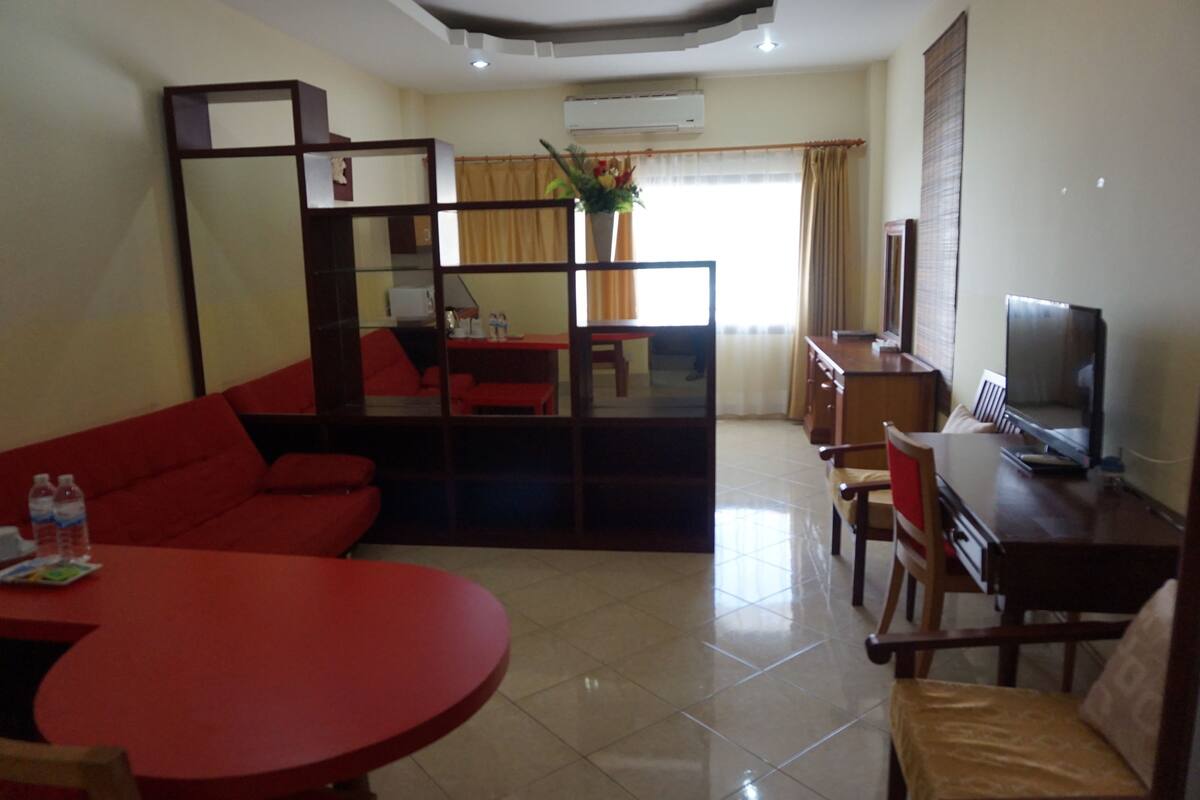 Ein Hotelzimmer mit einer roten Couch, einem Esstisch, einem Fernseher und einem großen Fenster mit Vorhängen.