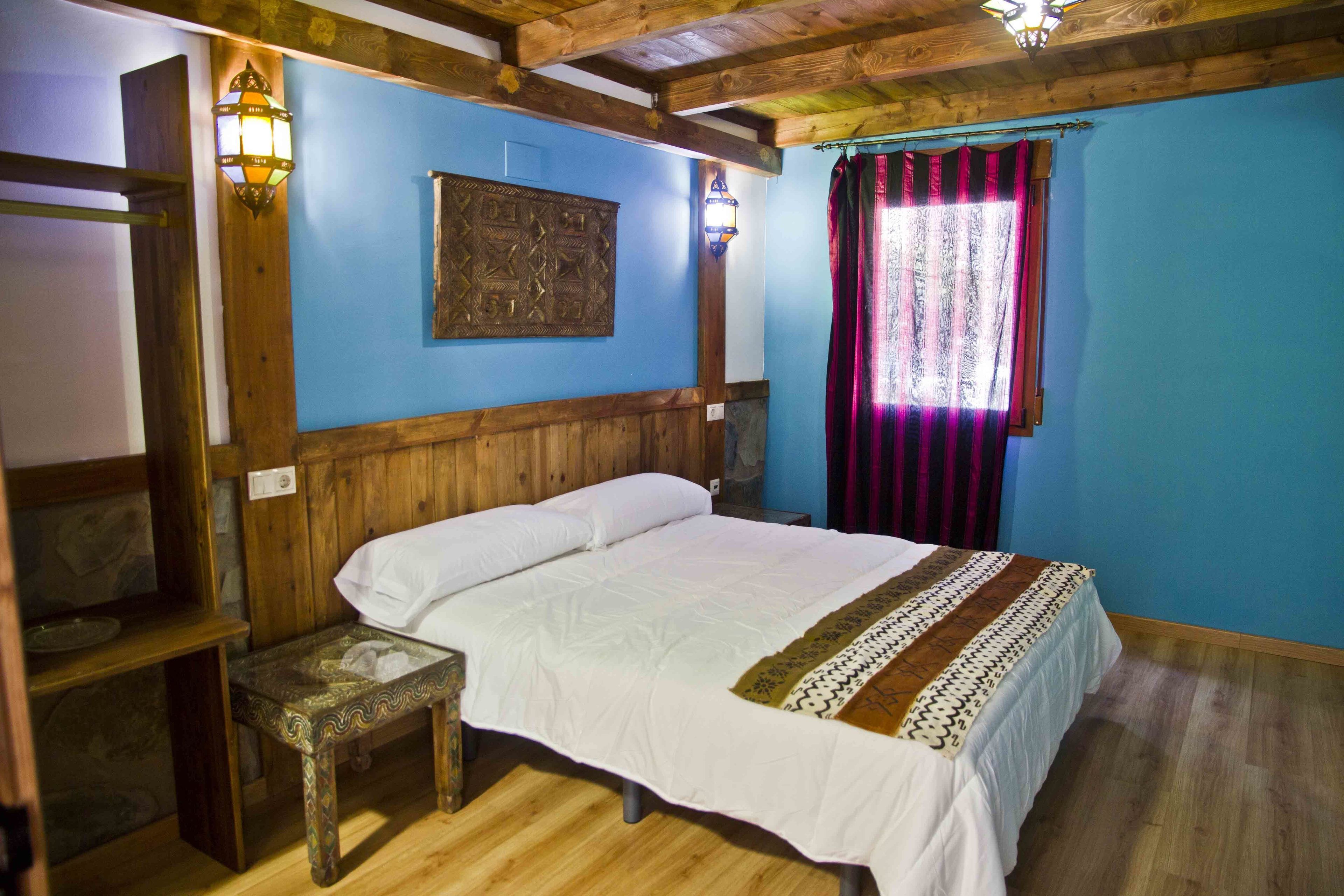 Una habitación con una cama de madera, dos lámparas fijadas a la pared, un tapiz decorado y una ventana con cortina roja.