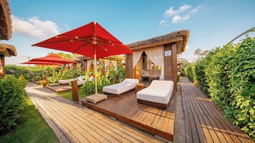 A wooden deck with sun loungers and red umbrellas, surrounded by lush greenery.