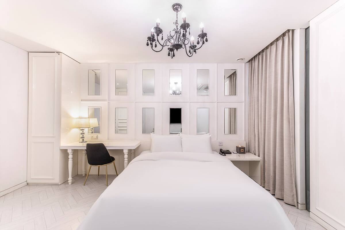 A modern hotel room with a large bed, a desk with a chair, a chandelier, and a view of the city through the windows.