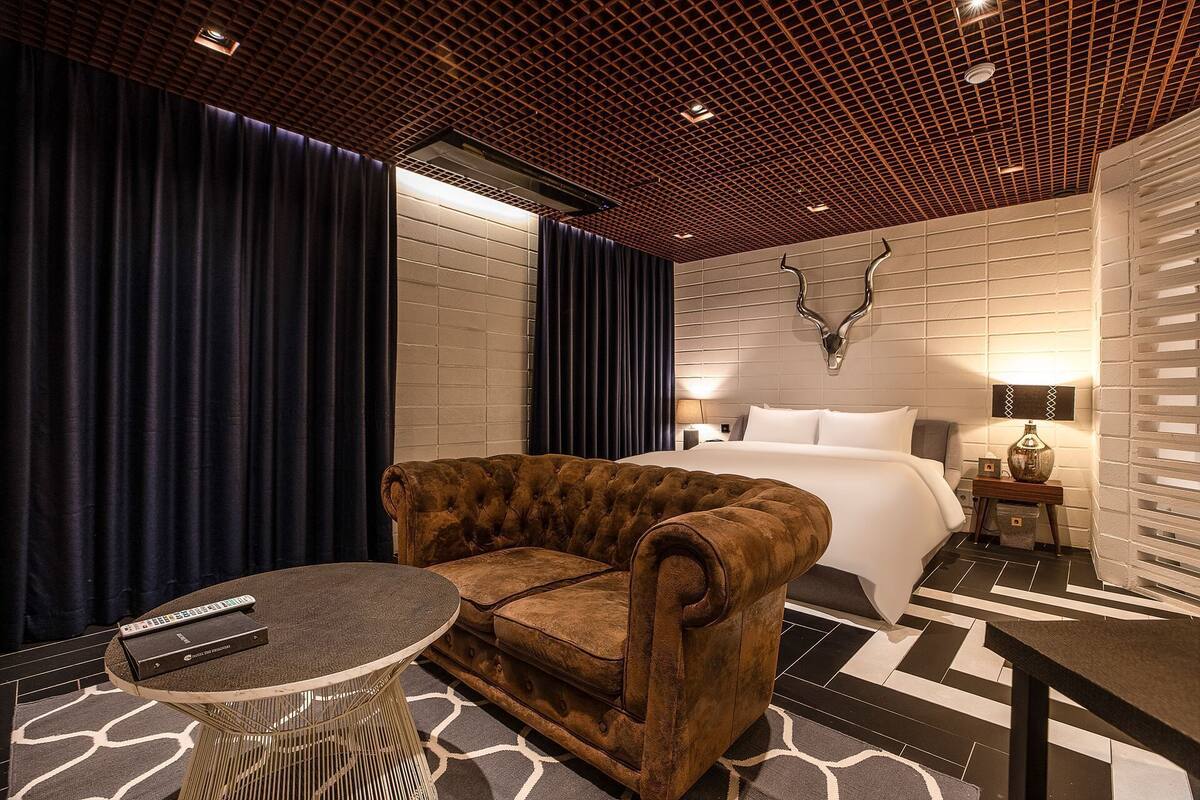 A hotel room with a tufted sofa, a round table, a bed with white linens, a wall-mounted TV, and a decorative wall piece.