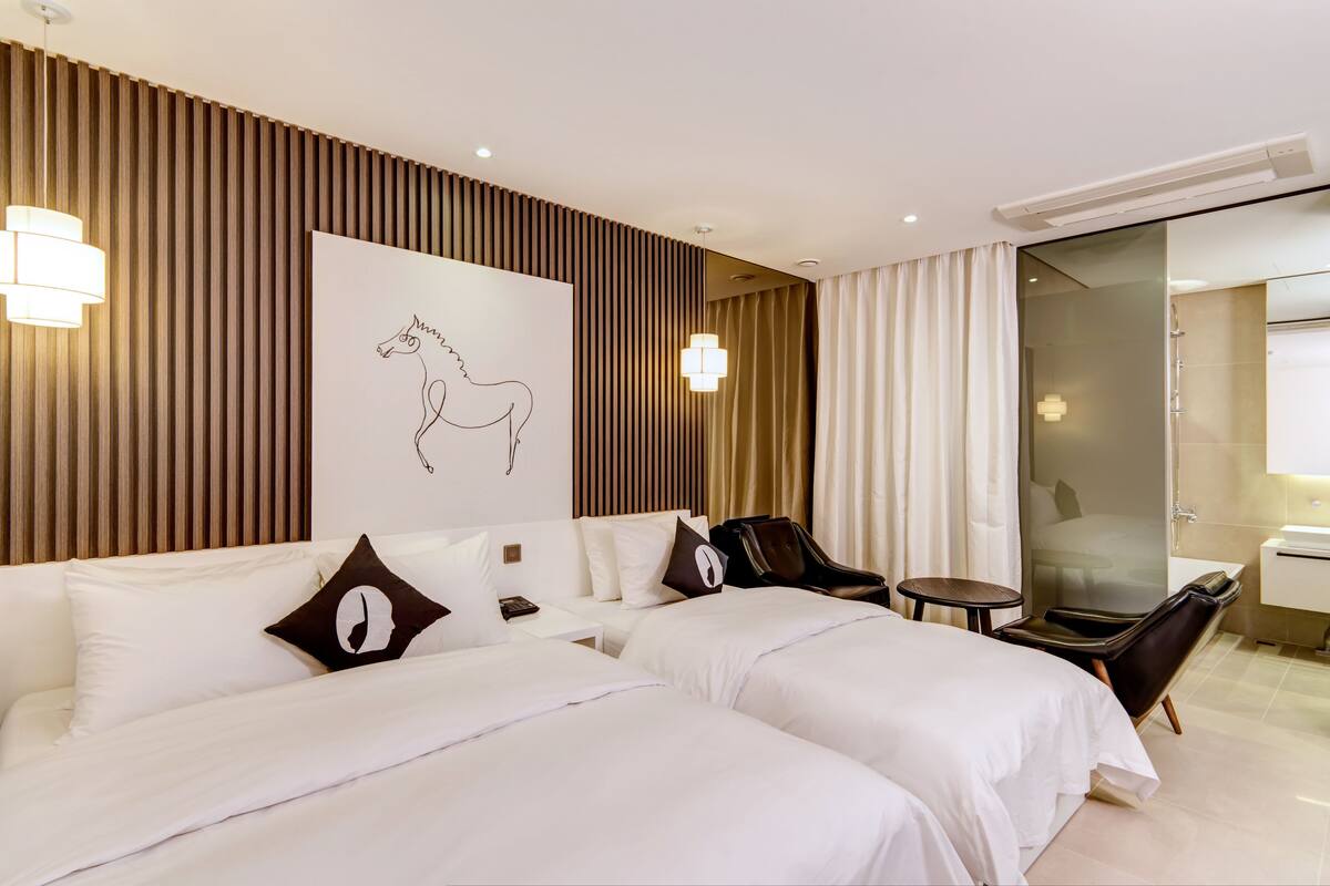 A hotel room with two beds, a large wall art of a horse, a seating area with a chair and table, and a glass shower enclosure.