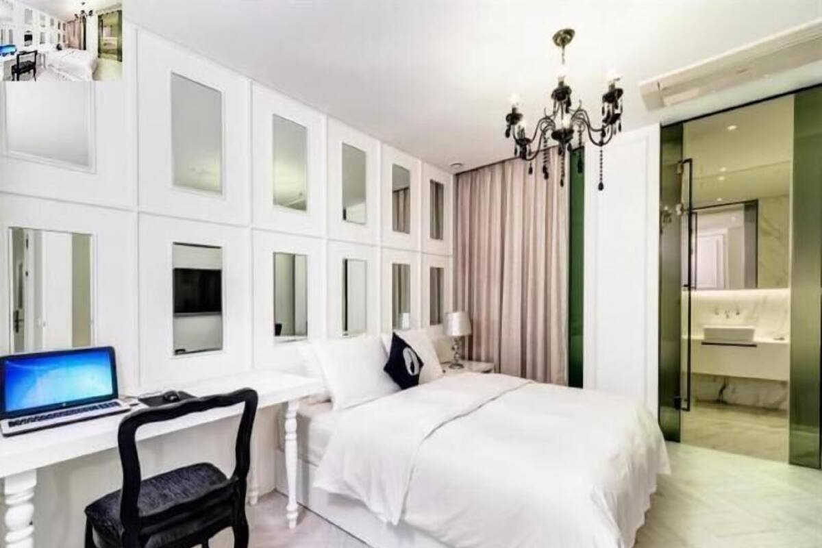 A hotel room with a white desk, a bed, a chair, a chandelier, and a bathroom with a mirror.