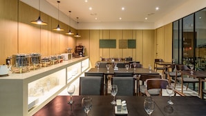 Un restaurante con paredes de paneles de madera, iluminación colgante y una barra con equipo para servicio de alimentos. Las mesas están preparadas con copas de vino y cubiertos.