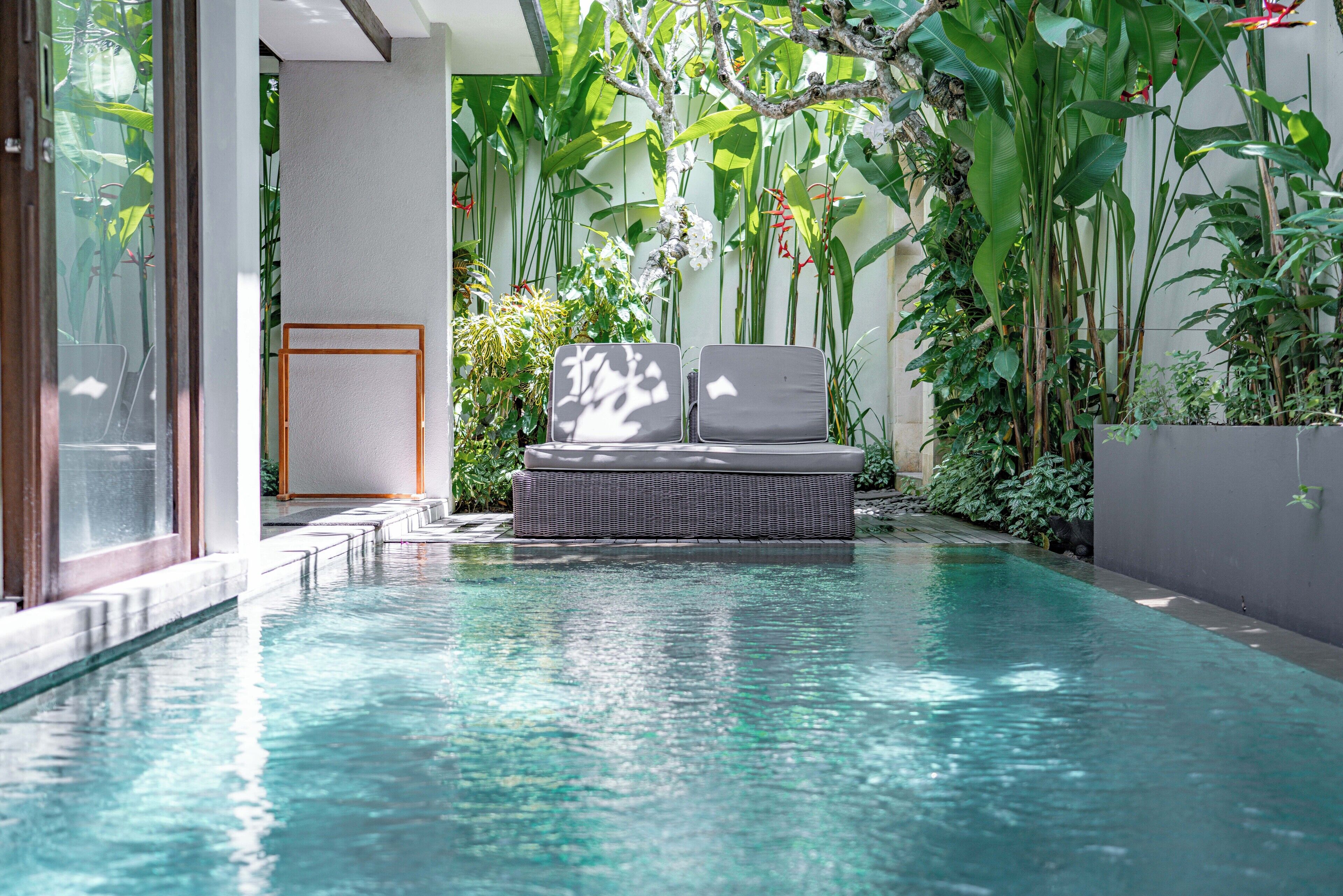 A pool area with a wicker lounge chair and a small table, surrounded by lush tropical plants.
