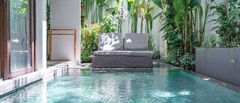 A pool area with a wicker lounge chair and a small table, surrounded by lush tropical plants.