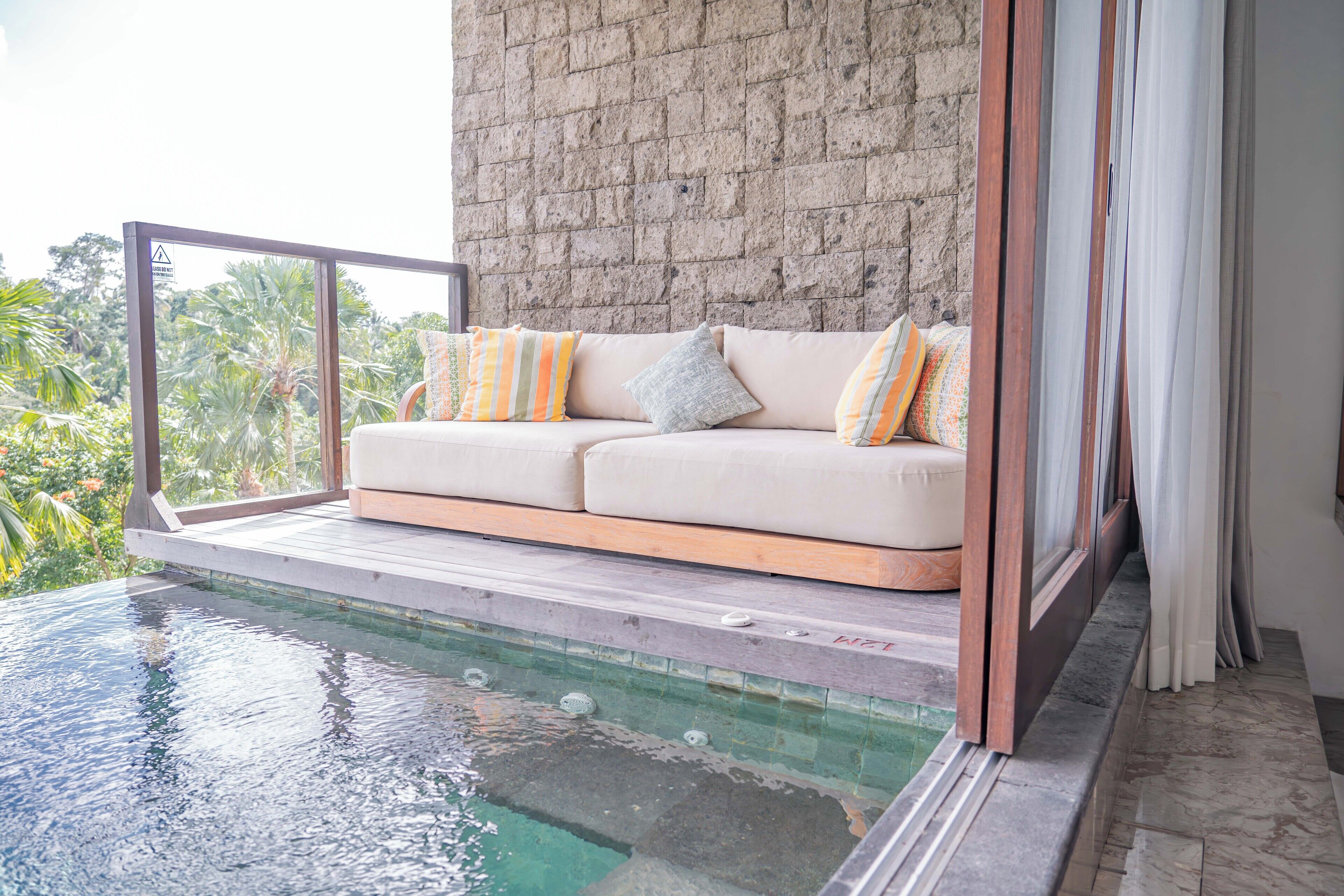 A balcony with a white sofa and cushions, a stone wall, and a pool.