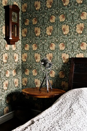 A room with floral wallpaper, a wooden side table with a lamp, and a bed with a patterned bedspread. - Okens Bed & Breakfast (Varberg)