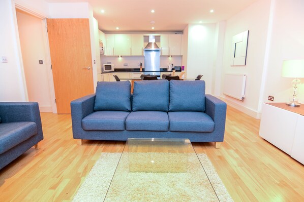 Ein modernes Wohnzimmer mit einem blauen Sofa, einem Glastisch und einer Küche im Hintergrund.